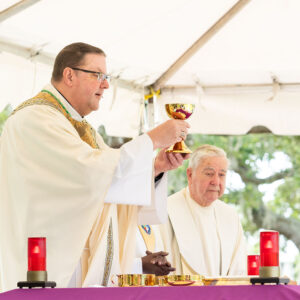 Bishop Parkes raises the chalice with the Most Precious Blood of Jesus Christ during the Consecration of the Mass on Memorial Day at Calvary Catholic Cemetery as Msgr. Michael Devine looks on at right.