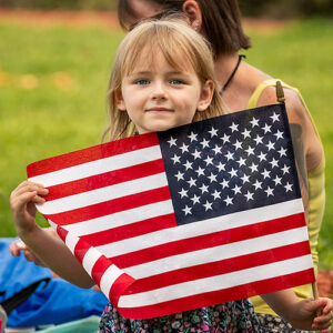 Four-year-old Ruby K. proudly displays a small American flag prior to Mass on Memorial Day at Calvary Catholic Cemetery. Her parents spread a blanket beneath the branches of a large oak tree in the park-like setting of the Cemetery.